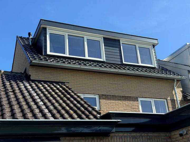 Dakkapel met witte kozijnen en antraciet zijwang op een pannendak, tegen een blauwe lucht